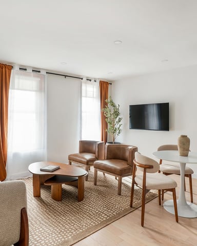 Open-plan living and dining area with beige seating, brown leather chairs, white tables, wall-mounted TV, orange curtains, and kitchen with stainless steel