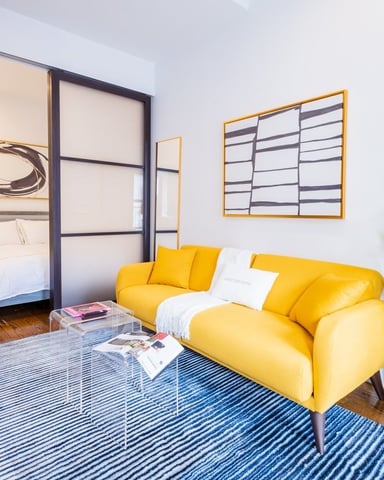 Studio apartment with yellow sofa, blue striped rug, and bedroom visible through glass partition door