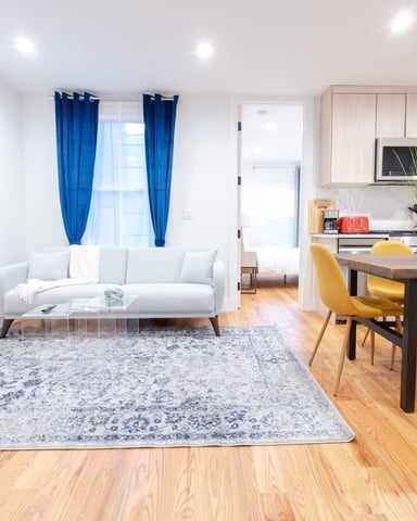 Open-plan living and dining area with light gray sofa, blue curtains, wooden dining table, yellow chairs, and modern kitchen with light wood cabinetry.