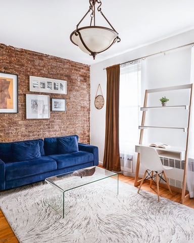 Living room with exposed brick wall, blue velvet sofa, glass coffee table, wooden shelving, and large windows with brown curtains