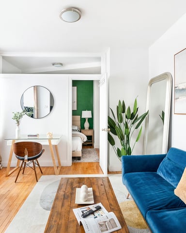 Bright open-plan living room with blue sofa, wooden TV stand, desk area, and bedroom visible through doorway with green accent wall