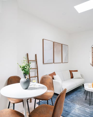 Bright living room with white sofa, round dining table, brown leather chairs, wooden window frame, and decorative mirrors on white walls.