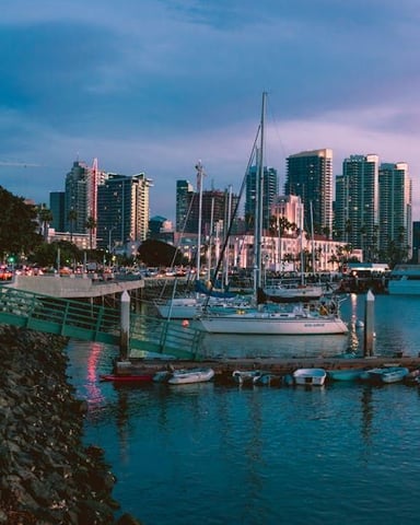 Waterfront promenade at dusk with sailboats, marina, and city skyline at twilight