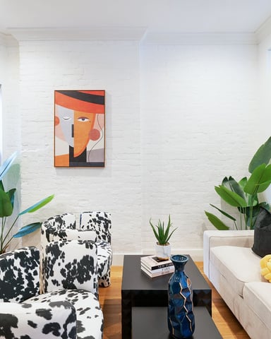 Bright living room with white brick walls, cow-print armchair, cream sofa with black pillows, potted plants, abstract orange artwork, and black-framed windows.