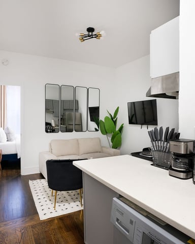 Modern studio kitchen with stainless steel appliances, white countertop, and open living area with beige sofa and mirrored wall panels