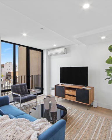 Modern living room with blue sofa, wood TV stand, large windows overlooking city, and potted plant
