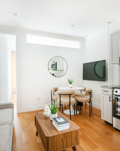 Modern open-plan living room and kitchen with white cabinetry, stainless steel appliances, wooden floors, and dining area with potted plants