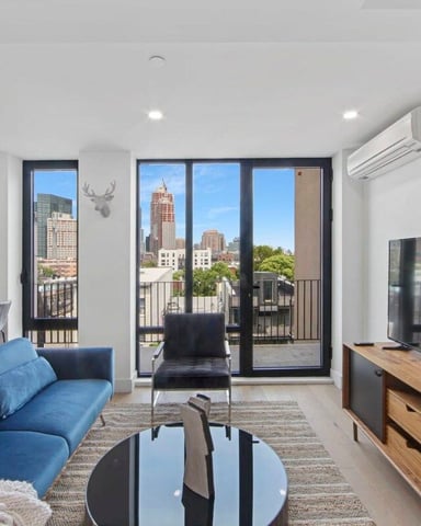 Modern living room with blue sofa, flat-screen TV, and floor-to-ceiling windows overlooking city skyline and tall buildings