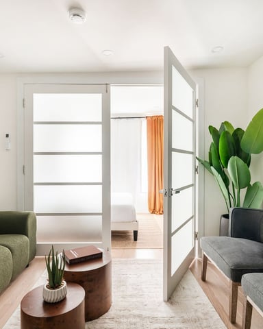 Modern living room with green curved sofa, gray seating, potted plant, wall-mounted TV, and glass doors opening to bedroom with orange curtains