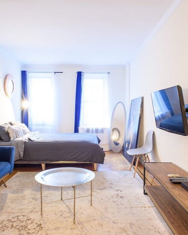 Bright studio bedroom with blue armchair, bed, wall-mounted TV, and two windows with blue curtains
