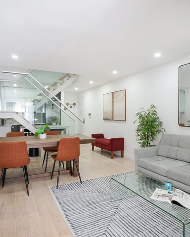 Modern open-plan living room with gray sofa, dining table with orange chairs, glass staircase, and wall-mounted mirrors