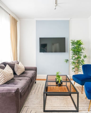 Modern living room with gray sofa, two blue armchairs, glass coffee table, wall-mounted TV, round mirror, and potted plant