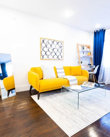 Bright living room with yellow sofa, glass coffee table, wooden floors, and blue curtains