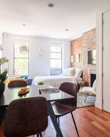 Studio apartment with dining table in foreground, bedroom with exposed brick wall and bed visible in background, large windows with natural light