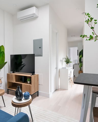 Modern open-plan living room with blue sofa, wooden TV stand, round mirror, potted plants, and dining table with black top.