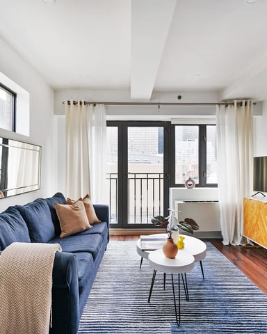 Modern studio apartment with blue sofa, white coffee table, hardwood floors, and balcony overlooking city buildings