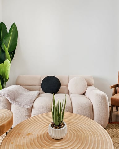 Modern living room with cream sofa, cognac leather chair, wooden side tables, potted plants, and large windows