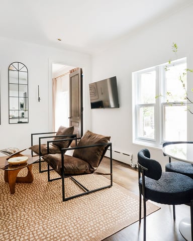 Bright open-plan living room with cream sofa, black leather chairs, arched mirrors, white walls, and potted greenery by glass doors
