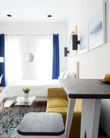 Studio apartment with exposed brick wall, bed with blue curtains, dining table with yellow chair, and hardwood floors