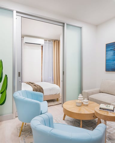 Modern studio with blue armchairs, beige sofa, wooden coffee tables, potted plant, and bedroom visible through glass doors