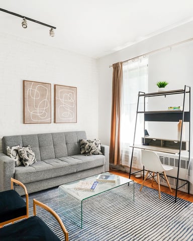 Bright living room with gray sofa, striped rug, metal shelving unit, and large windows with tan curtains