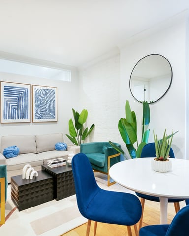 Bright living room with white walls, round dining table, blue velvet chairs, beige sofa, teal armchairs, potted plants, and abstract blue artwork.