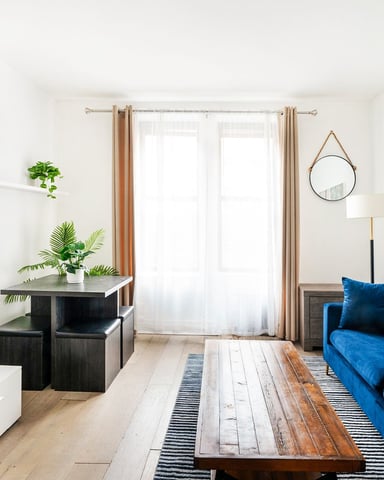 Modern living room with blue sofa, wall-mounted TV, wooden coffee table, potted plants, and glass doors opening to natural light