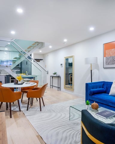 Modern open-plan living space with blue sofa, orange dining chairs, glass staircase, and abstract wall art