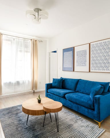 Bright living room with blue velvet sofa, wooden coffee table, framed wall art, and glass doors with cream curtains
