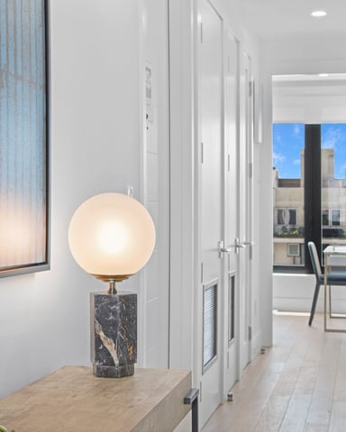 Modern hallway with white walls, marble table lamp, tall cactus plant, and view to balcony with city skyline