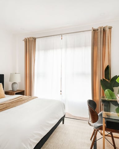 Modern bedroom with black upholstered bed, tan curtains, desk with plants, and wall-mounted TV