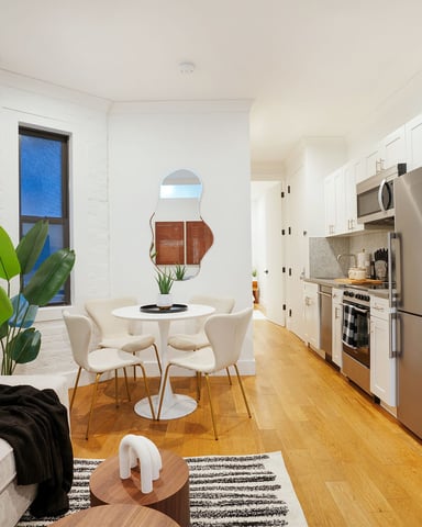 Open-plan kitchen and dining area with white brick walls, round dining table, cream chairs, stainless steel appliances, and hardwood floors.