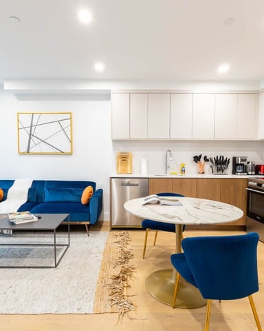 Modern open-plan apartment with blue sofa, marble dining table, wood and white kitchen, and floor-to-ceiling windows overlooking city.