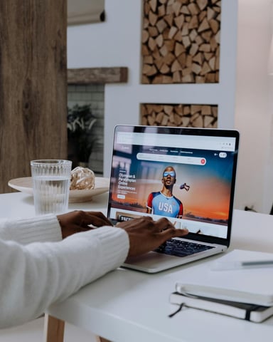 Person at white desk using laptop displaying sports website, with glass of water, notebooks, and floor lamp nearby