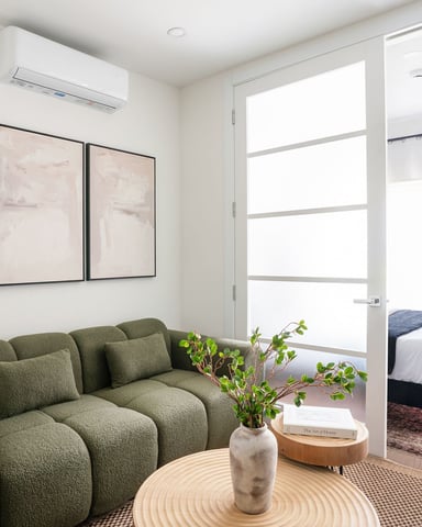 Modern studio with olive-green sofa, round wood table, three framed abstract artworks, and bedroom visible through glass door