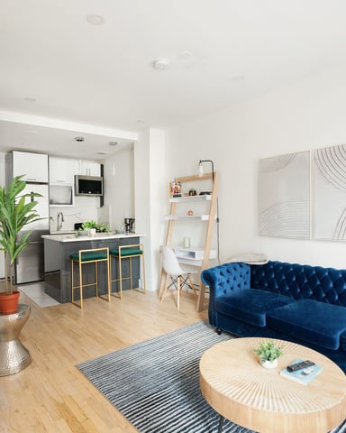 Open-plan studio with blue velvet sofa, wooden coffee table, and kitchen with gray island in background