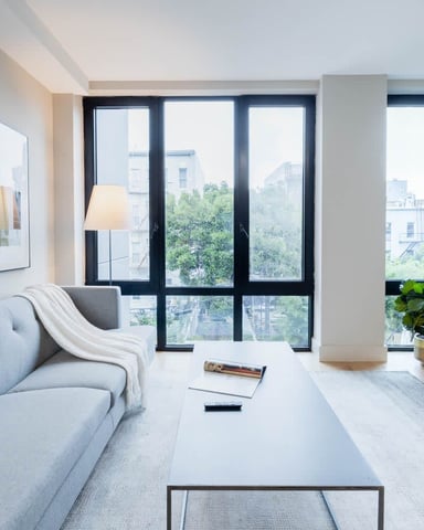 Modern living room with gray sofa, floor-to-ceiling windows overlooking city buildings, TV on dark wood console, and potted plant.