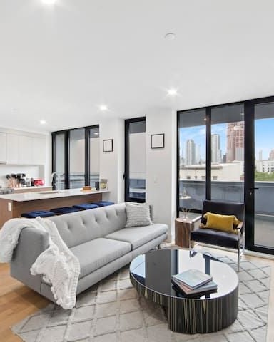 Open-plan living room and kitchen with gray sofa, round coffee table, and floor-to-ceiling windows overlooking city skyline