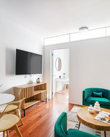 Modern living room with wall-mounted TV, wooden furniture, emerald green chairs, hardwood floors, and potted plants throughout