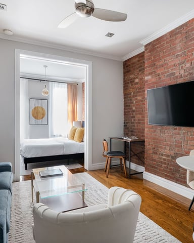 Studio apartment with exposed brick wall, TV, blue sofa, white armchair, and bedroom visible through doorway