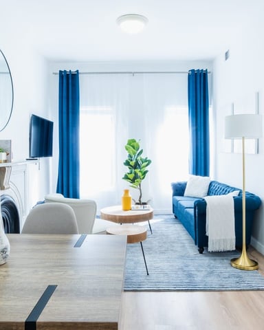 Bright modern living room with blue sofa, blue curtains, round mirror, and white ceramic vases on wooden floors