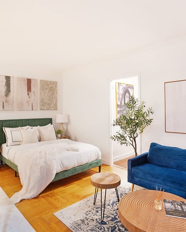 Studio bedroom with green upholstered bed, blue sofa, wooden desk, potted plant, and hardwood floors