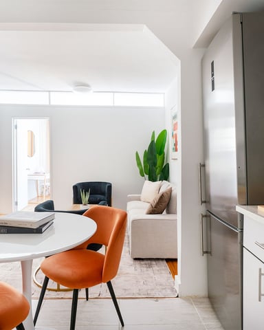 Modern kitchen with white cabinetry and stainless steel appliances opening to living room with orange chairs and round white table