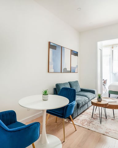 Modern living room with blue velvet chairs, white round table, gray sofa, potted plant, and bedroom visible through open doorway