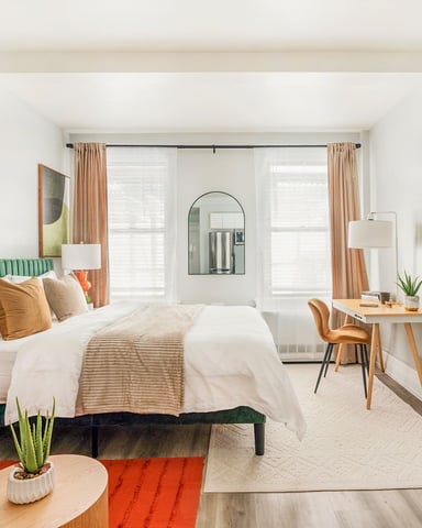 Modern studio bedroom with green headboard, white bedding, desk, seating area, and floor-to-ceiling windows with tan curtains