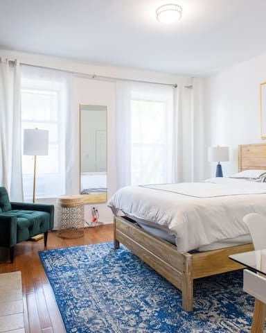 Bright bedroom with wooden bed, white bedding, blue patterned rug, green armchair, and framed abstract art above headboard