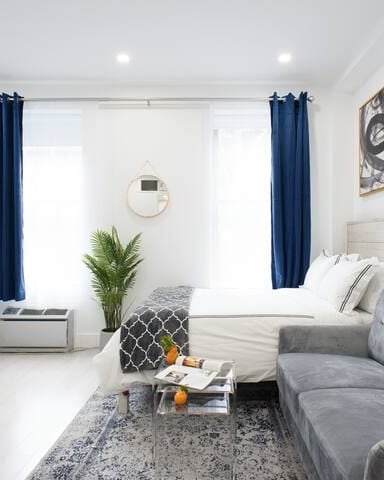 Bright studio apartment with gray sofa, white bed, desk, large windows with navy curtains, and gold wall decor