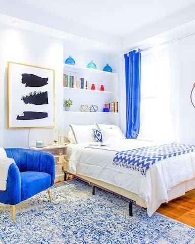 Bright bedroom with white walls, blue accent chair, white bed with blue patterned throw, floating shelves, and wooden flooring