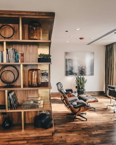 Modern office with wooden shelving unit, lounge chairs, desk, and floor-to-ceiling windows overlooking city buildings