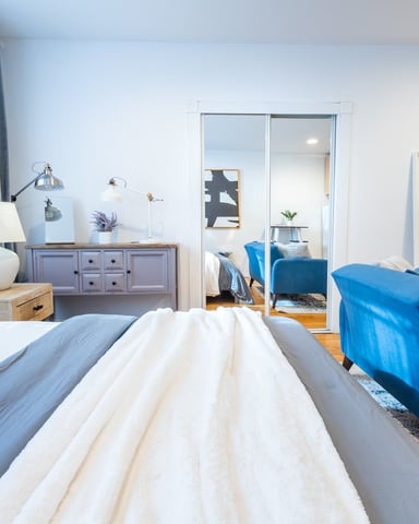 Bright bedroom with white walls, blue bedding, mirrored closet doors, and sitting area with blue armchair and wall-mounted TV